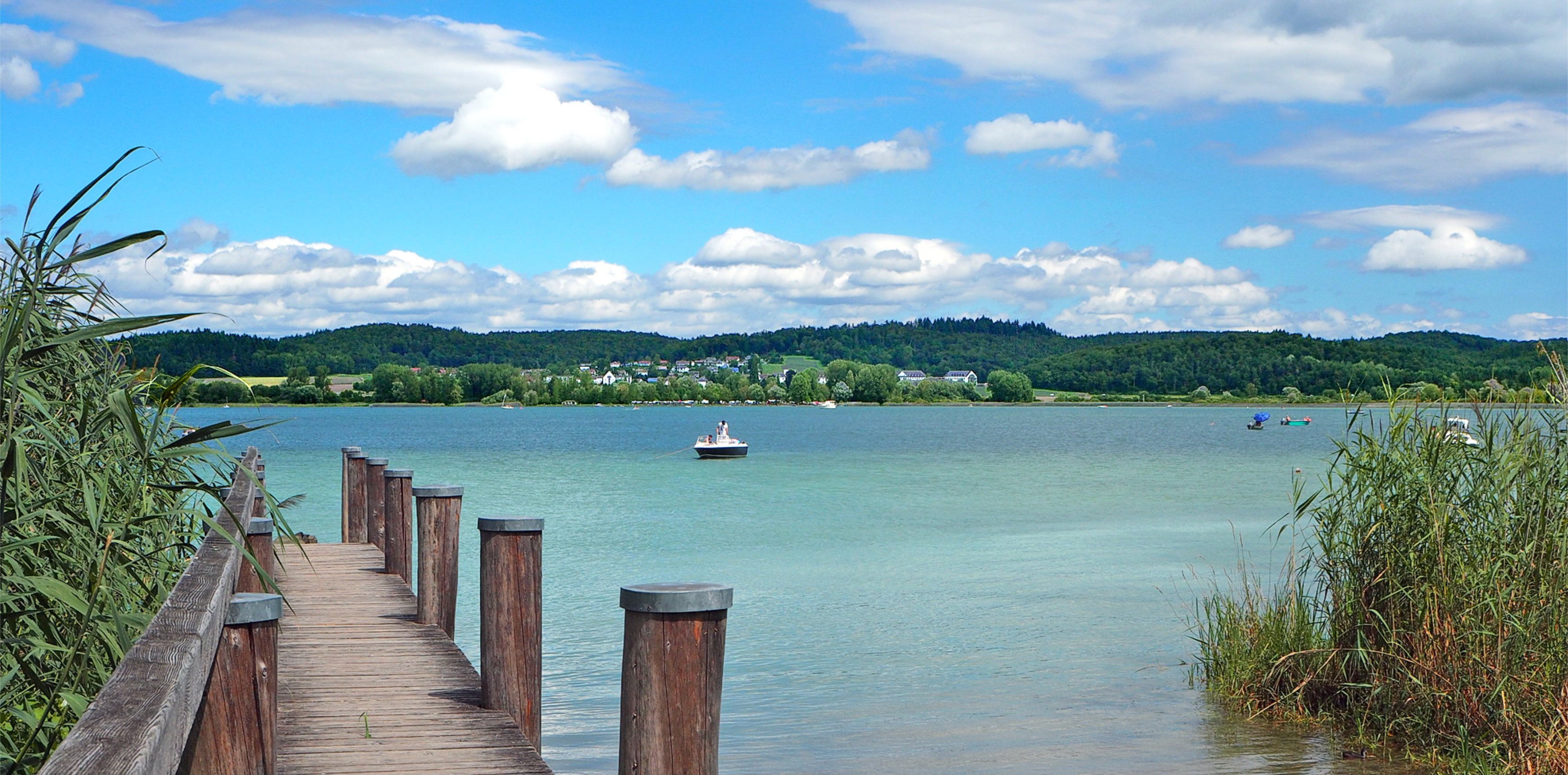Holzsteg führt durch Schilf in einen See; ein Motorboot liegt im Wasser, dahinter liegen Dorf, bewaldete Hügel und Wolkenhimmel.