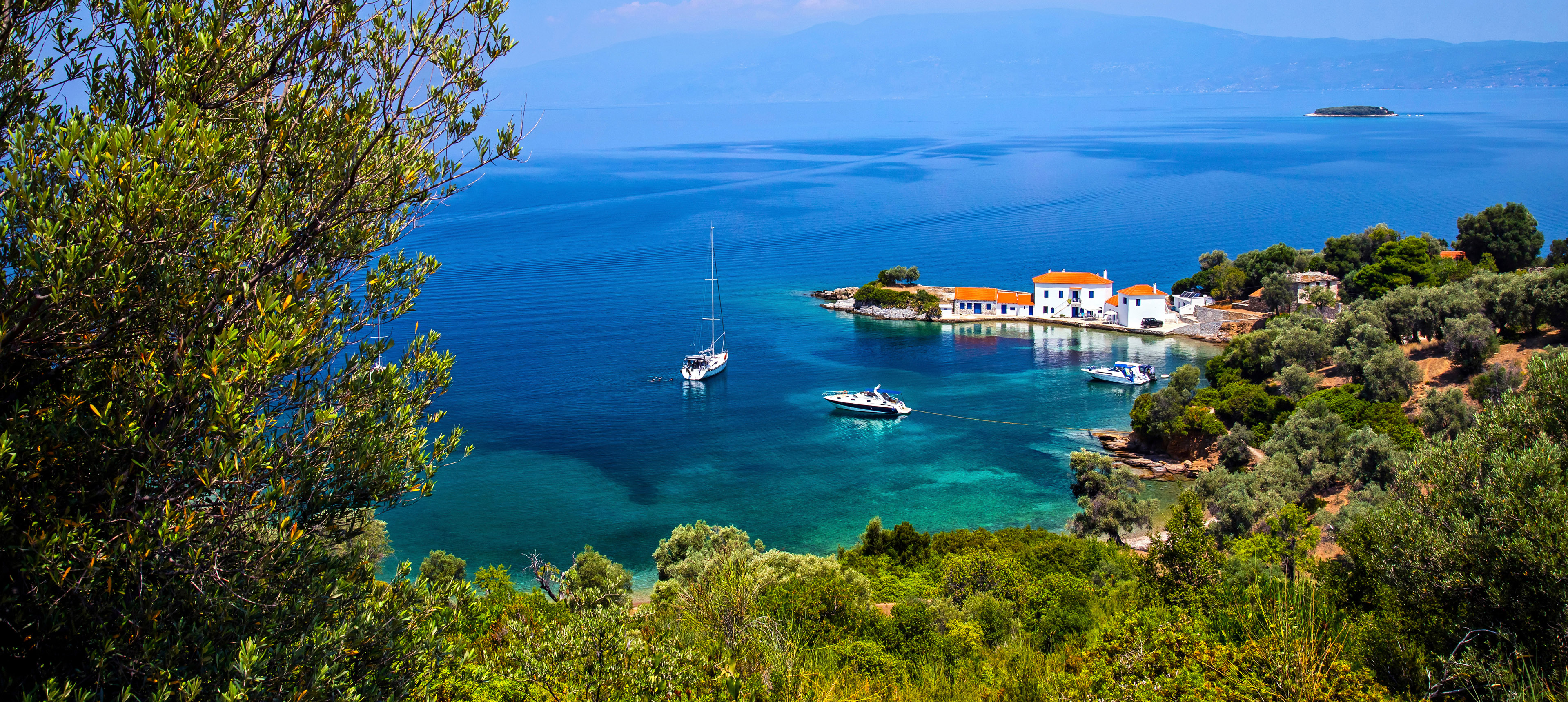 Griechische Küstenlandschaft mit weißen Häusern, roten Dächern, Olivenbäumen und Booten im türkisblauen Meer.