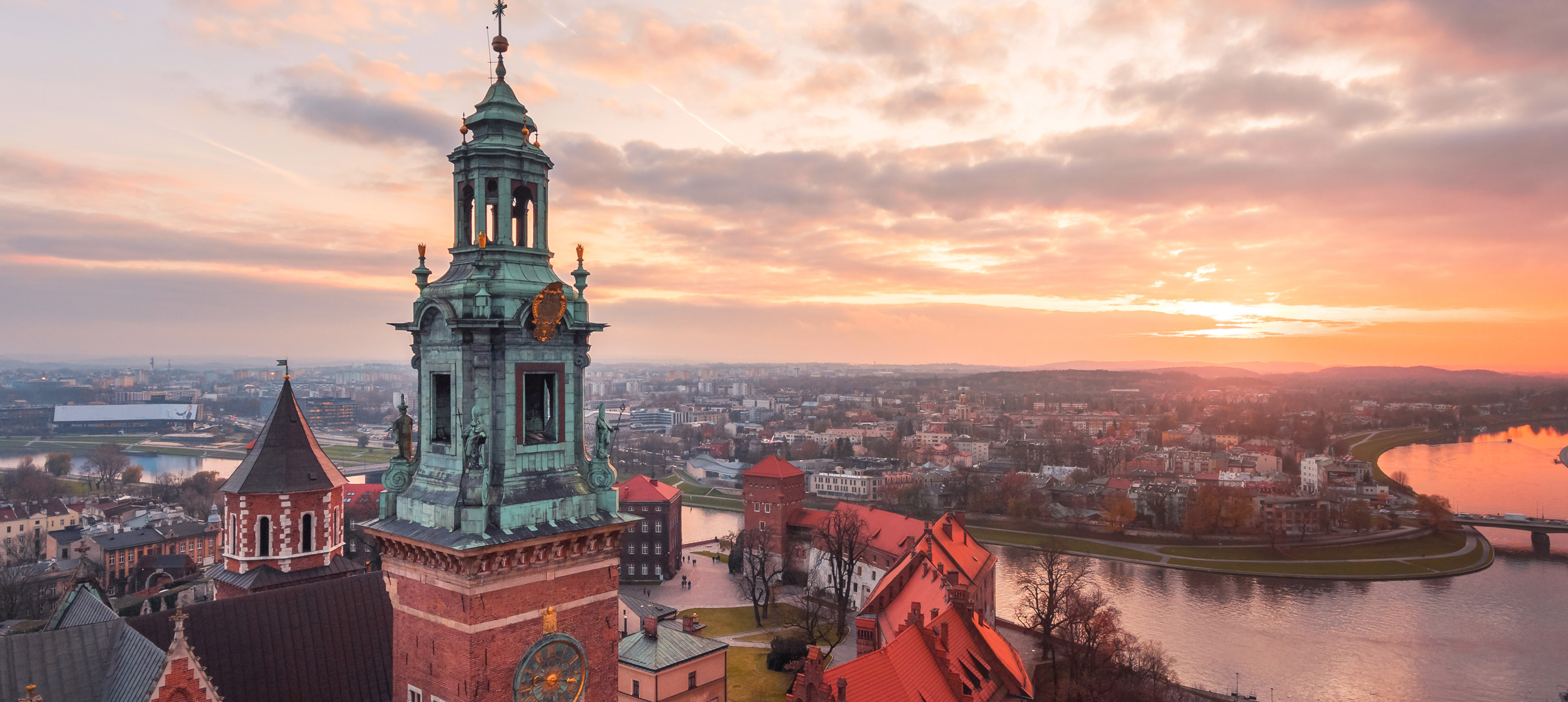 Das Bild zeigt den Wasserturm der Wawel-Kathedrale in Krakau bei Sonnenuntergang. Im Hintergrund verläuft der Fluss Wisła, umgeben von historischen und modernen Gebäuden. Die warmen Farben des Himmels verleihen der Szene eine ruhige, malerische Atmosphäre.