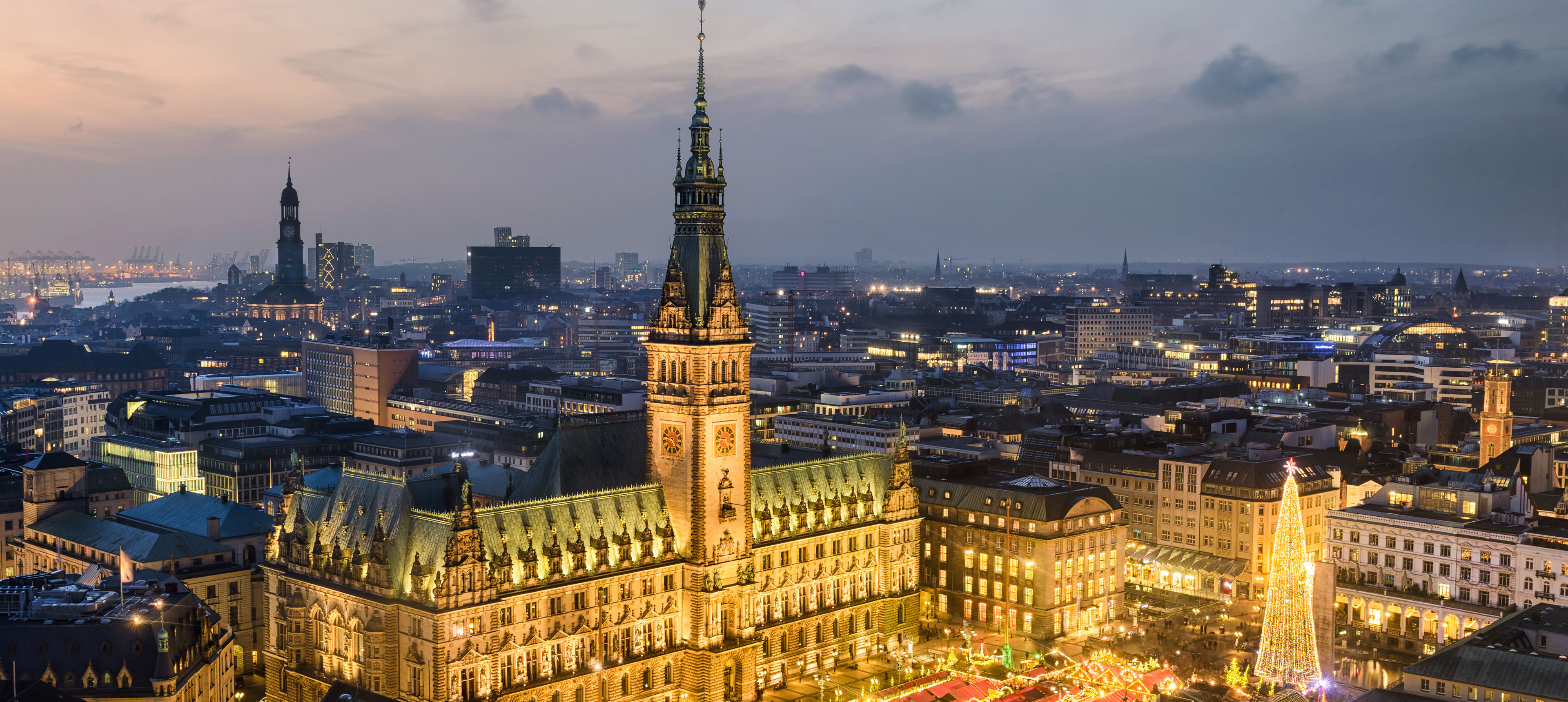 Abendliche Panoramaaufnahme des festlich beleuchteten Hamburger Rathauses, umgeben von der erleuchteten Innenstadt. Im Vordergrund ein großer Weihnachtsmarkt mit Lichtern und Tannenbaum, im Hintergrund Hafenkräne und die Silhouette der Stadt.