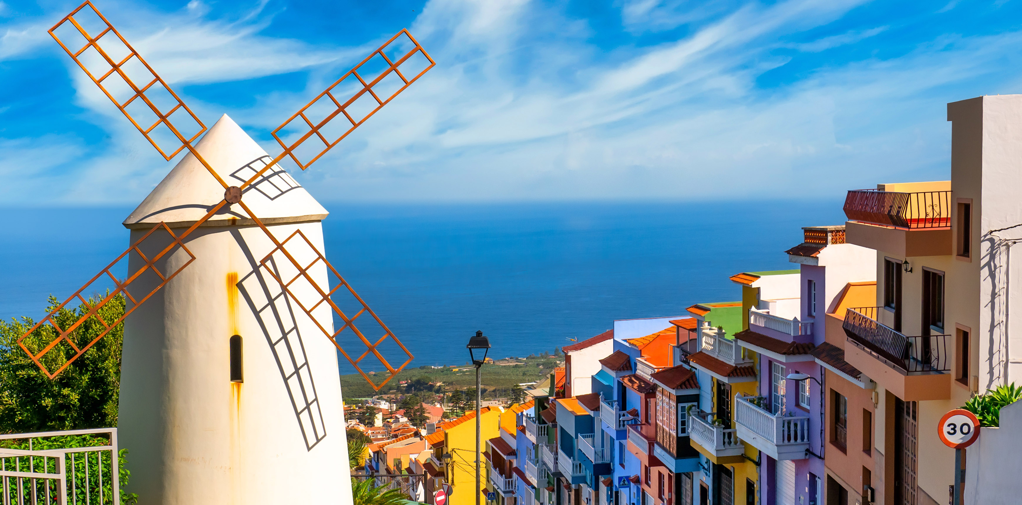 Weiße Windmühle mit orangefarbenen Flügeln über einer bunten Küstenstadt mit Blick auf das blaue Meer.