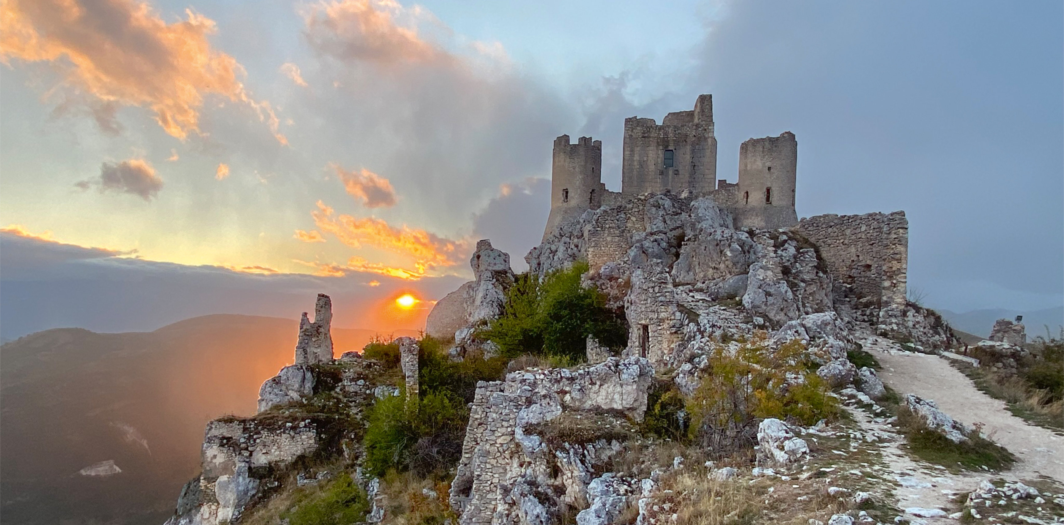 Burgruine auf Felsen im Sonnenuntergang, mit steinigem Weg und orangefarbenem Himmel.