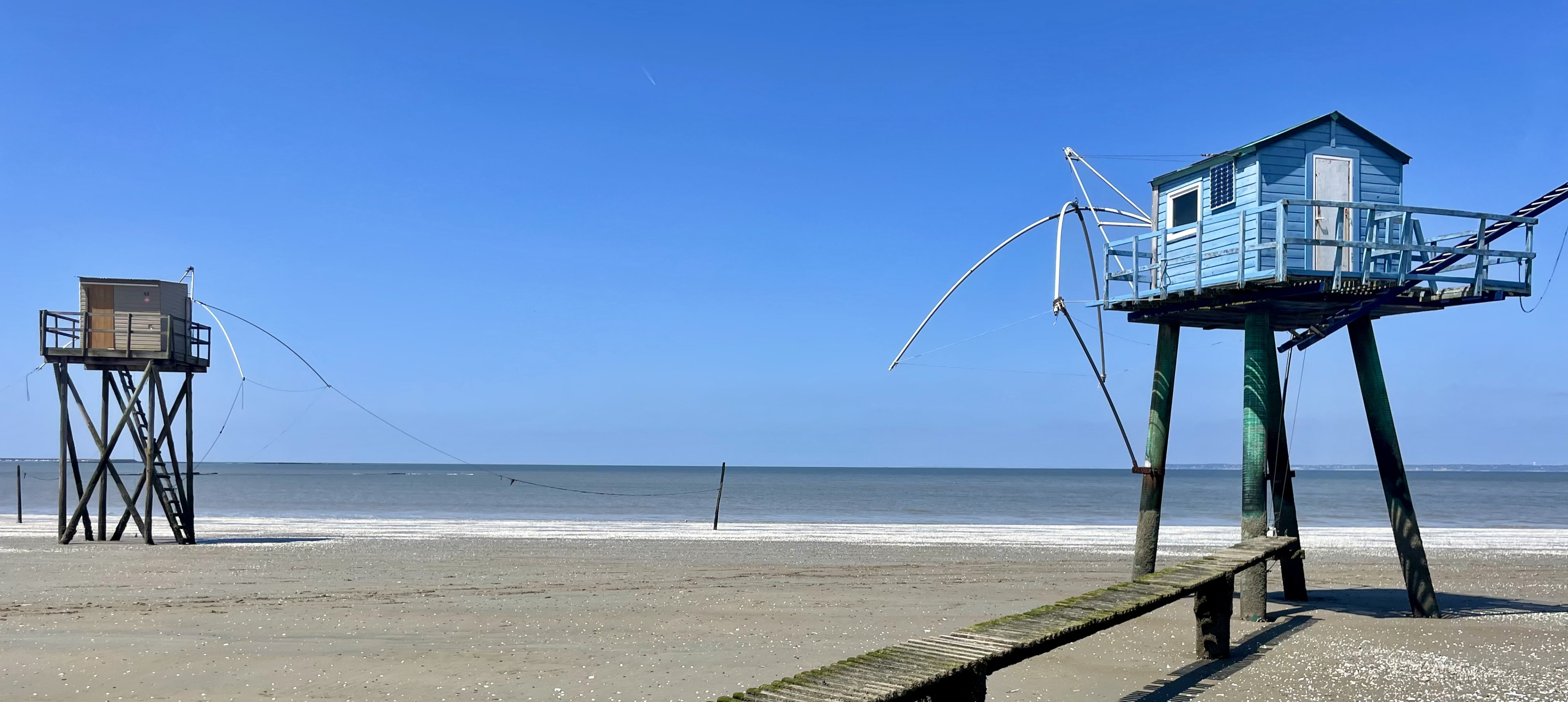 Zwei Stelzenfischerhütten an einem flachen Sandstrand unter klarem blauen Himmel.