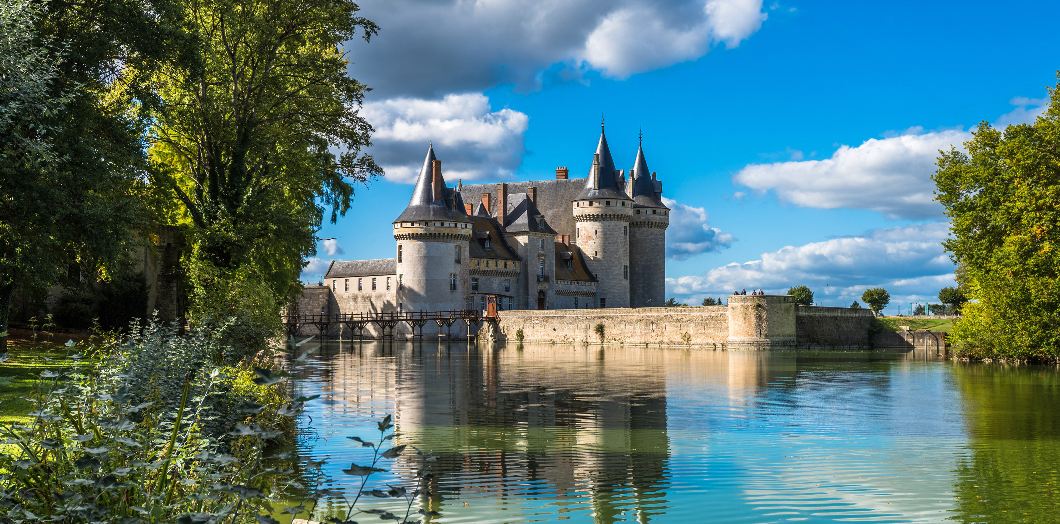 Märchenhaftes Schloss mit Türmen und Zinnen, davor ein ruhiger Fluss, in dem sich das Schloss spiegelt, eingebettet in eine grüne Landschaft unter blauem Himmel mit weißen Wolken.