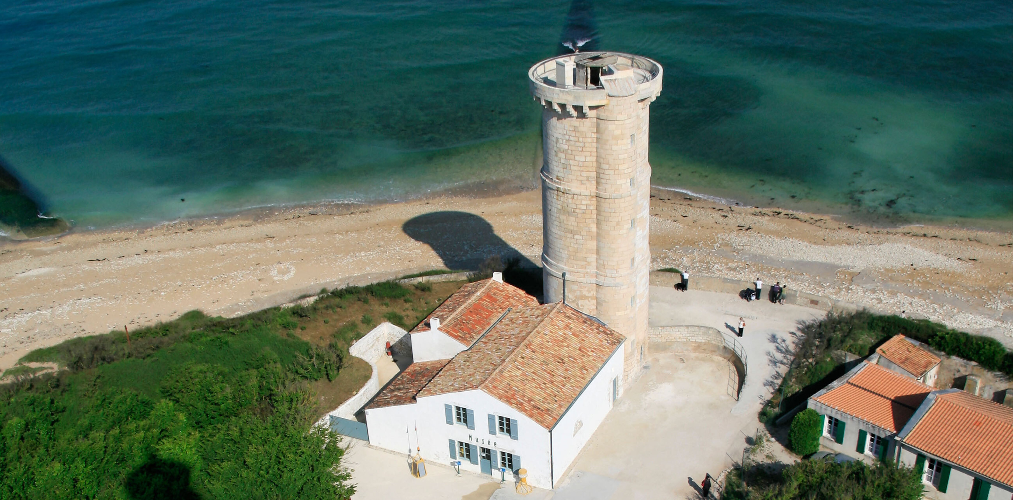 Steinturm am Strand neben weißem Museumshaus mit rotem Ziegeldach und türkisfarbenem Meer.