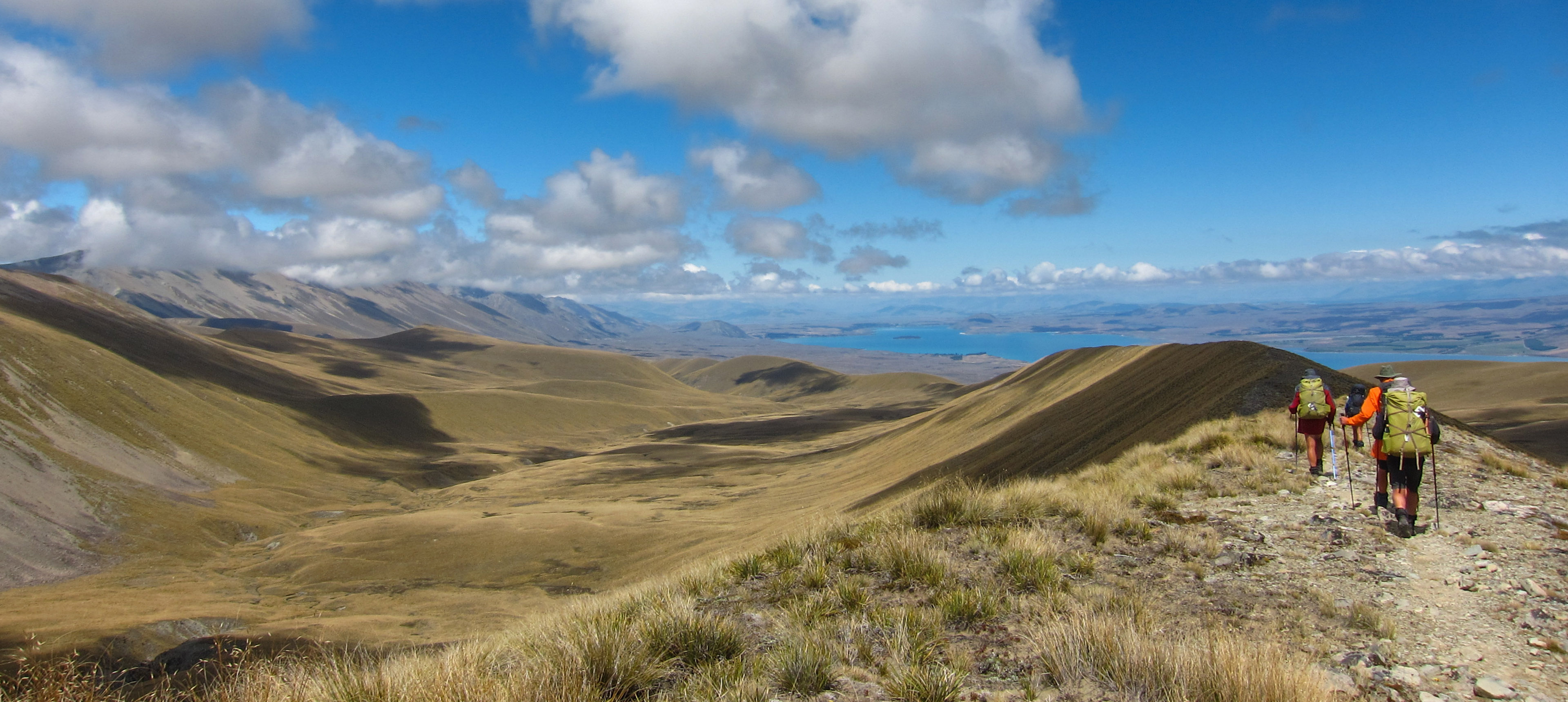Wanderer mit Rucksäcken auf einem Bergpfad in weiter, goldbrauner Hügellandschaft unter blauem Himmel mit weißen Wolken – Blick über Täler und in der Ferne schimmernde Seen.