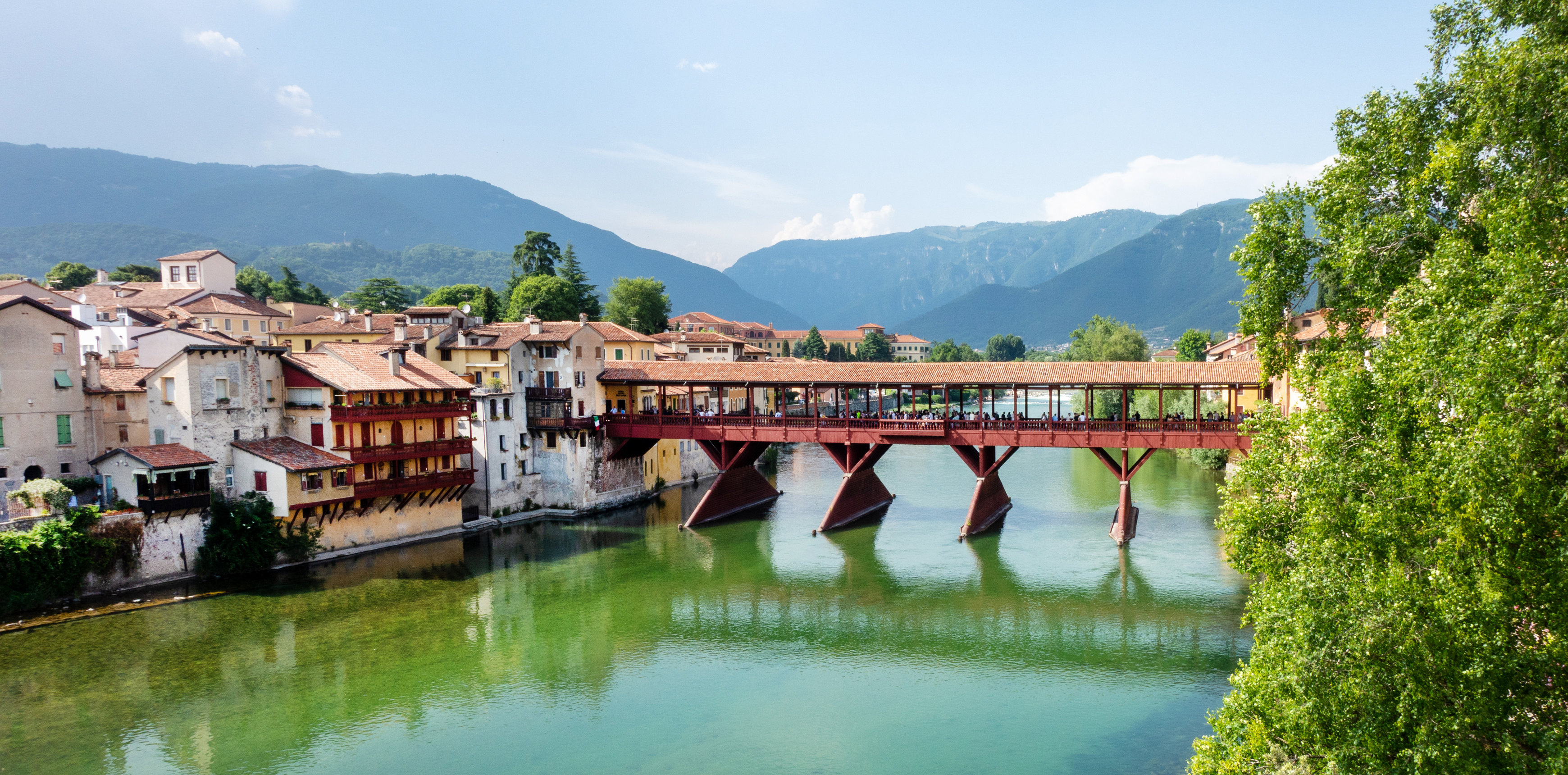 Holzüberdachte Brücke Ponte degli Alpini in Bassano del Grappa über den Fluss Brenta, umgeben von historischen Häusern, grünen Ufern und Bergen im Hintergrund unter blauem Himmel.