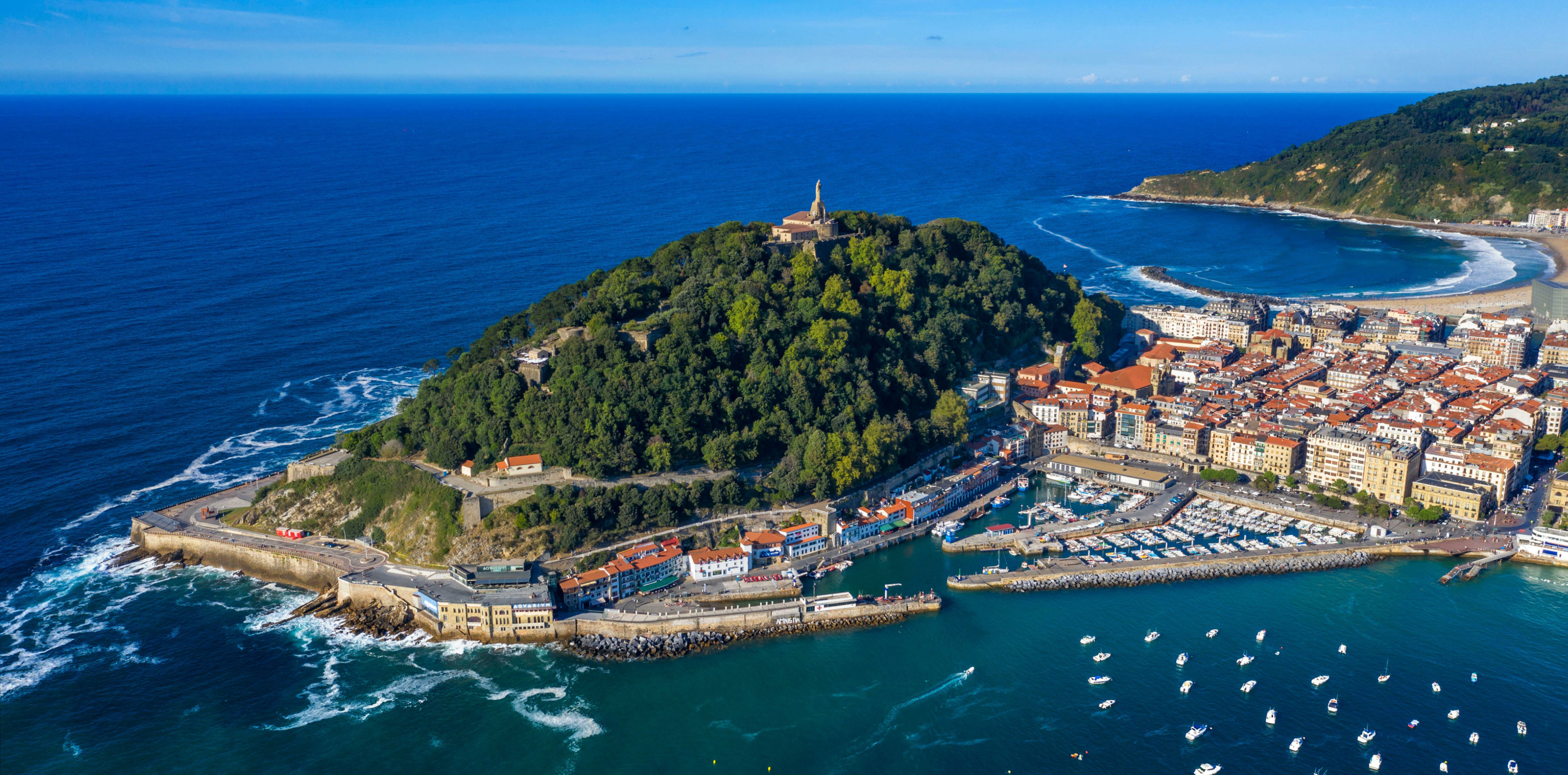 Luftaufnahme von San Sebastián mit bewaldetem Hügel, Kirche, Altstadt, Hafen, Stränden und zahlreichen Booten vor der Küste.