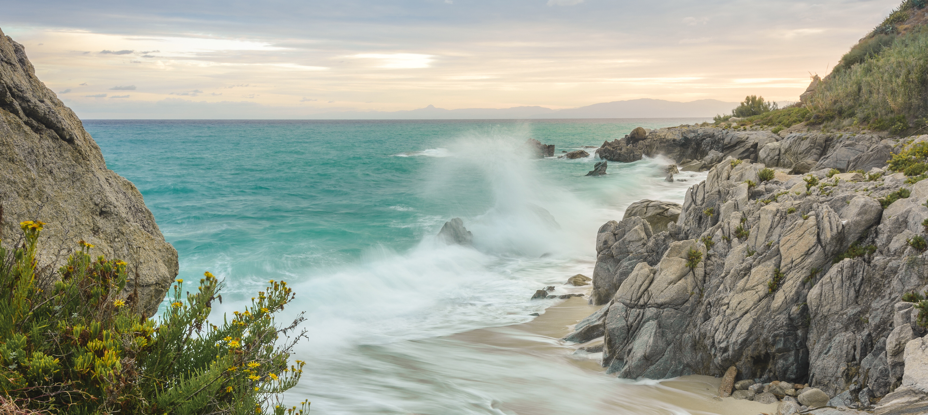Türkisfarbenes Meer mit weißer Gischt an einer felsigen Küste, einem schmalen Sandstrand und gelb blühenden Pflanzen im Vordergrund.