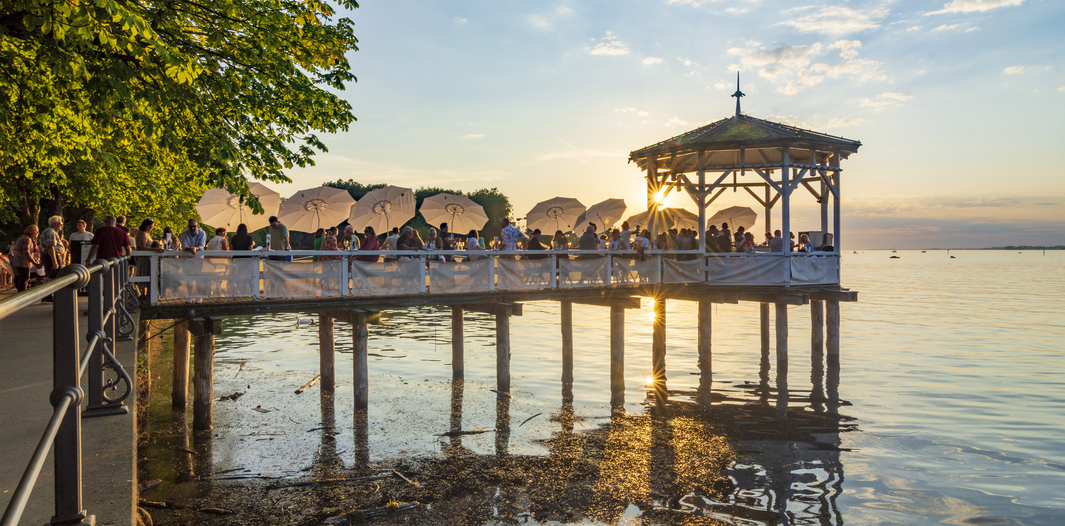 Menschen auf einer Seeterrasse bei Sonnenuntergang, viele weiße Sonnenschirme und ein Pavillon auf Pfählen im Wasser.